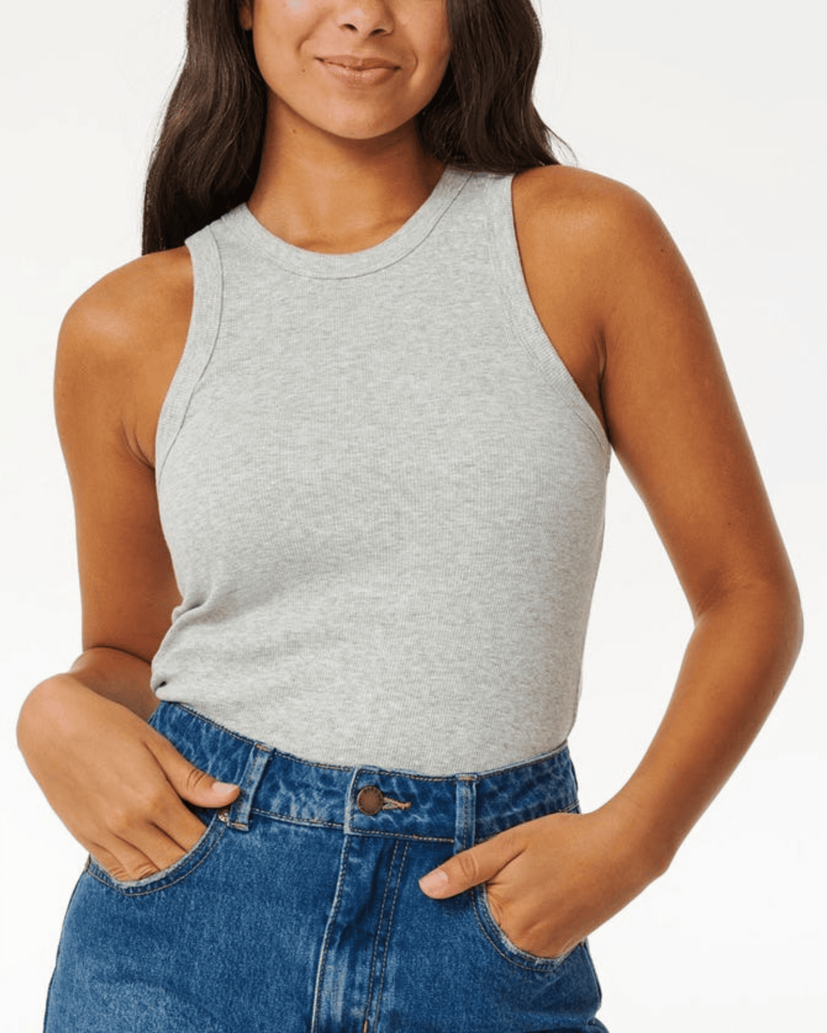 A person with long dark hair wears the Rip Curl Classic Ribbed Tank in gray and high-waisted blue jeans made of cotton elastane, standing with hands partially in pockets against a plain white background.