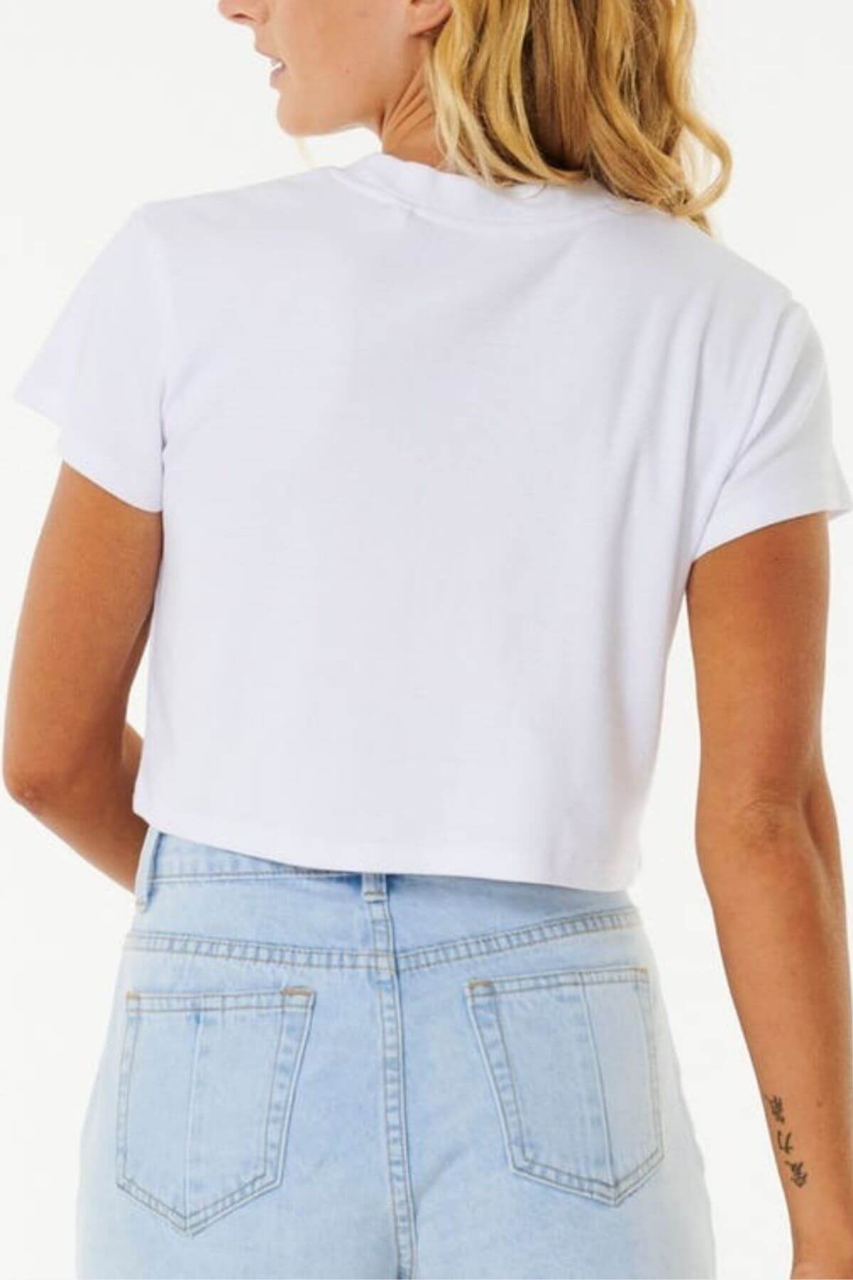 A person with blonde hair, viewed from behind, wears a Rip Curl Classic Ribbed Tee in white and light blue high-waisted jeans. A small tattoo on their left arm adds intrigue, while the plain white background highlights this wardrobe staple.