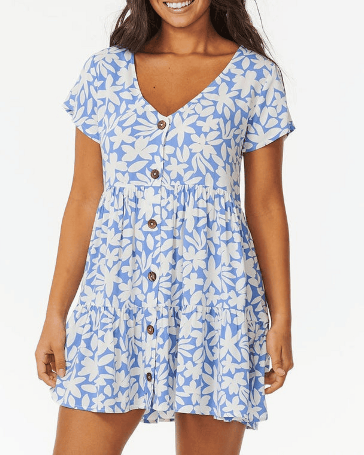 A woman smiles against a white background, wearing the Rip Curl Holiday Tropics Dress—a short-sleeved, light blue dress with white floral print, coconut front buttons, and a V-neckline.