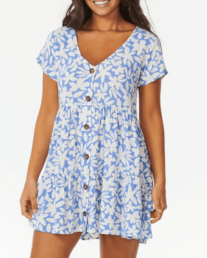 A woman smiles against a white background, wearing the Rip Curl Holiday Tropics Dress—a short-sleeved, light blue dress with white floral print, coconut front buttons, and a V-neckline.