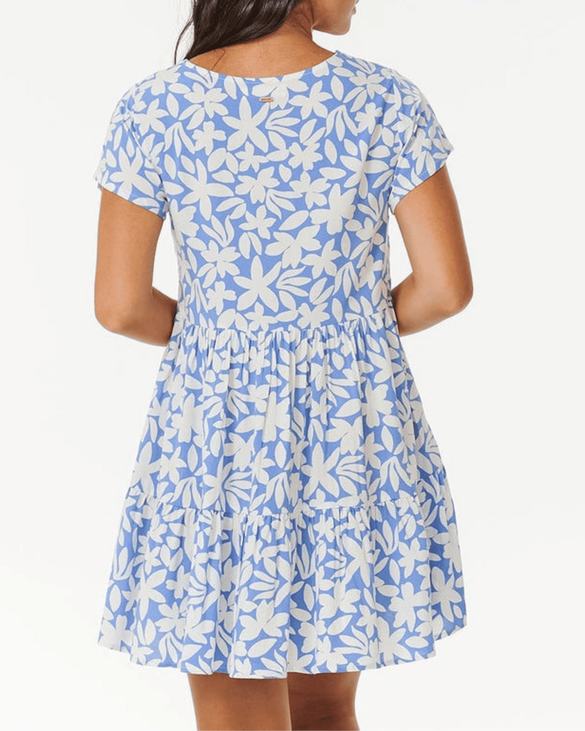 A woman is seen from the back wearing the Rip Curl Holiday Tropics Dress, which features short sleeves, a blue color with white yardage print, and a loose, tiered ruffled skirt.
