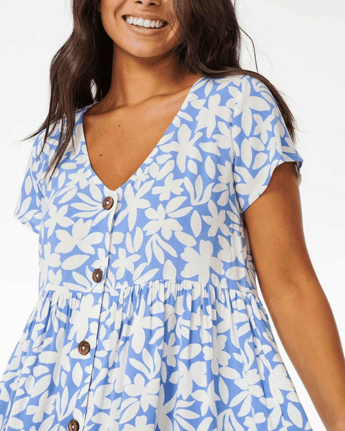 Smiling woman wears the Rip Curl Holiday Tropics Dress, a light blue dress with white floral print, short sleeves, V-neckline, and brown coconut buttons down the front.