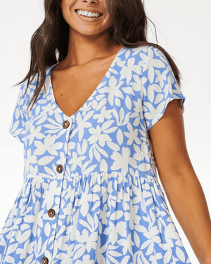 Smiling woman wears the Rip Curl Holiday Tropics Dress, a light blue dress with white floral print, short sleeves, V-neckline, and brown coconut buttons down the front.