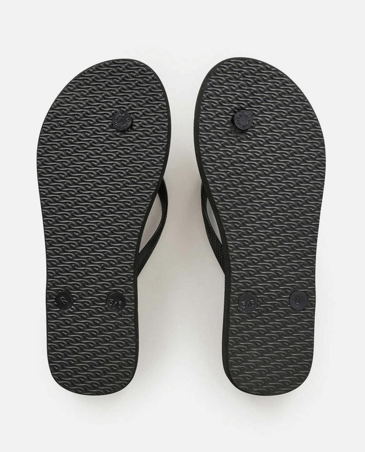A pair of Rip Curl La Isla Bloom Thongs, in black with textured soles, is displayed with the soles facing up. The right flip-flop shows noticeable wear on the heel area, while the left one appears less worn. Made with BLOOM™ algae blended foam for a sustainable touch, they are set against a plain white background.