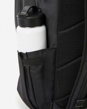 The close-up image highlights the Rip Curl Overtime 30L Backpack, made from recycled polyester, featuring a side mesh pocket with a white water bottle inside, and designed with a textured padded back and adjustable straps for comfort.