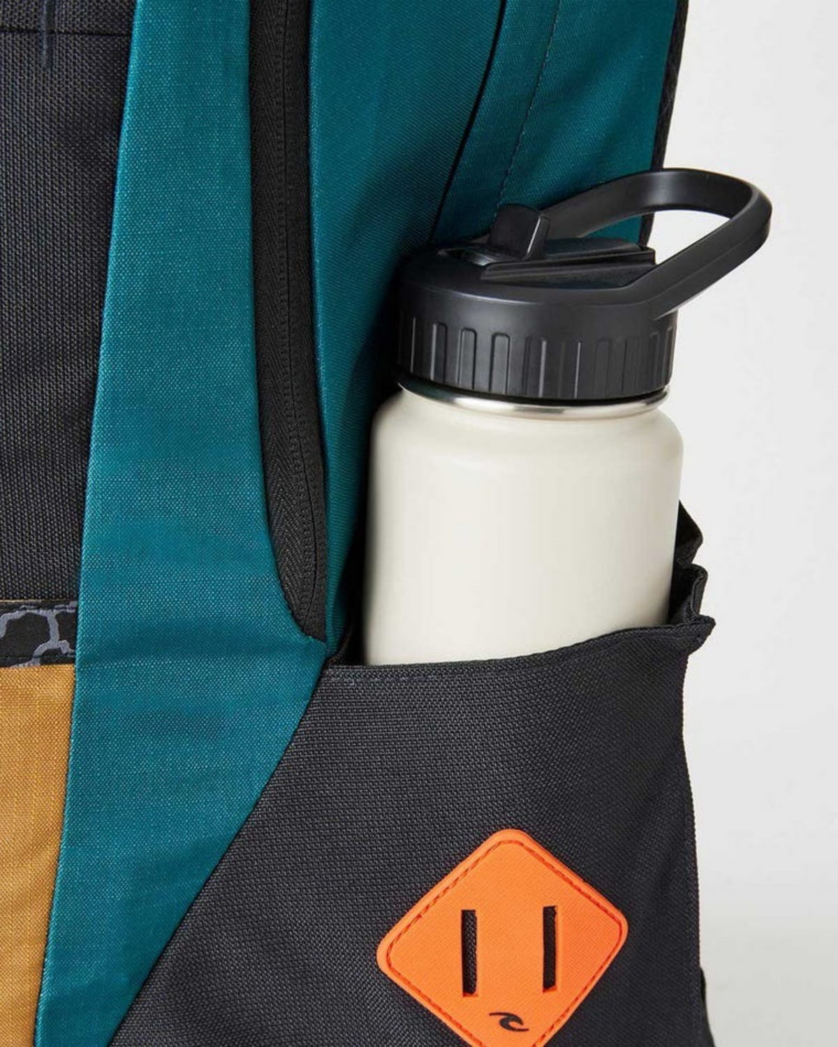A white water bottle with a black lid sits in the side pocket of the eco-friendly Rip Curl Overtime 30L Journeys Backpack, which features teal, black, and brown fabric and an orange diamond-shaped patch.