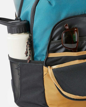 Close-up of a Rip Curl Ozone 30L Journeys Backpack with a blue top and tan pocket made from recycled polyester. A white water bottle sits in the side mesh pocket, while brown-framed sunglasses are tucked into the front. The backpack features black zippers and a rope zipper pull.