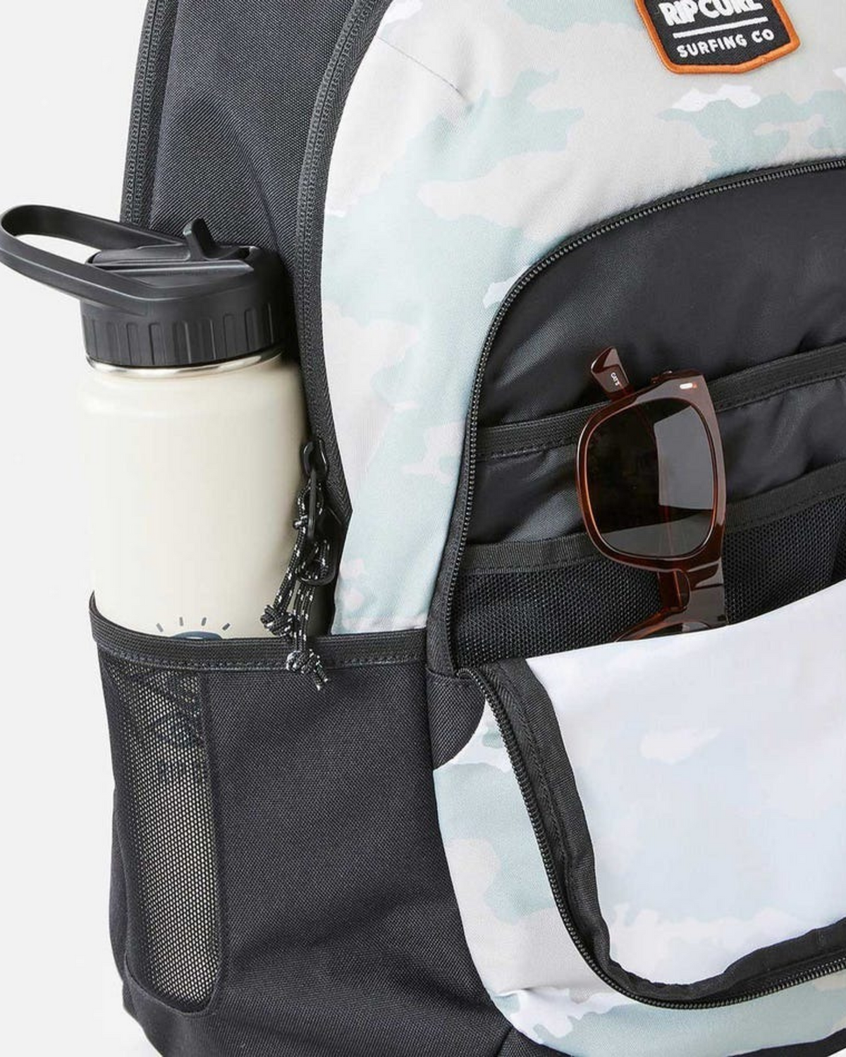 A close-up of the Rip Curl Ozone 30L School Backpack, made from recycled polyester with a light camouflage pattern. A white water bottle is in the side mesh pocket, and brown sunglasses are tucked into the front zip pocket.