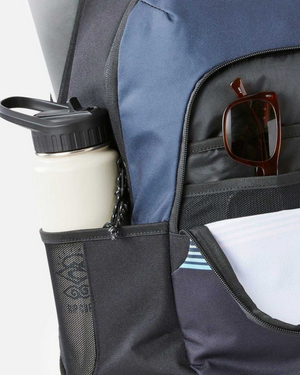 A close-up of the Rip Curl Ozone 30L School Backpack reveals a navy blue and black design with side pockets holding a white water bottle, front pockets for sunglasses, visible zippers, and mesh detailing. Crafted from recycled polyester for an eco-conscious style.