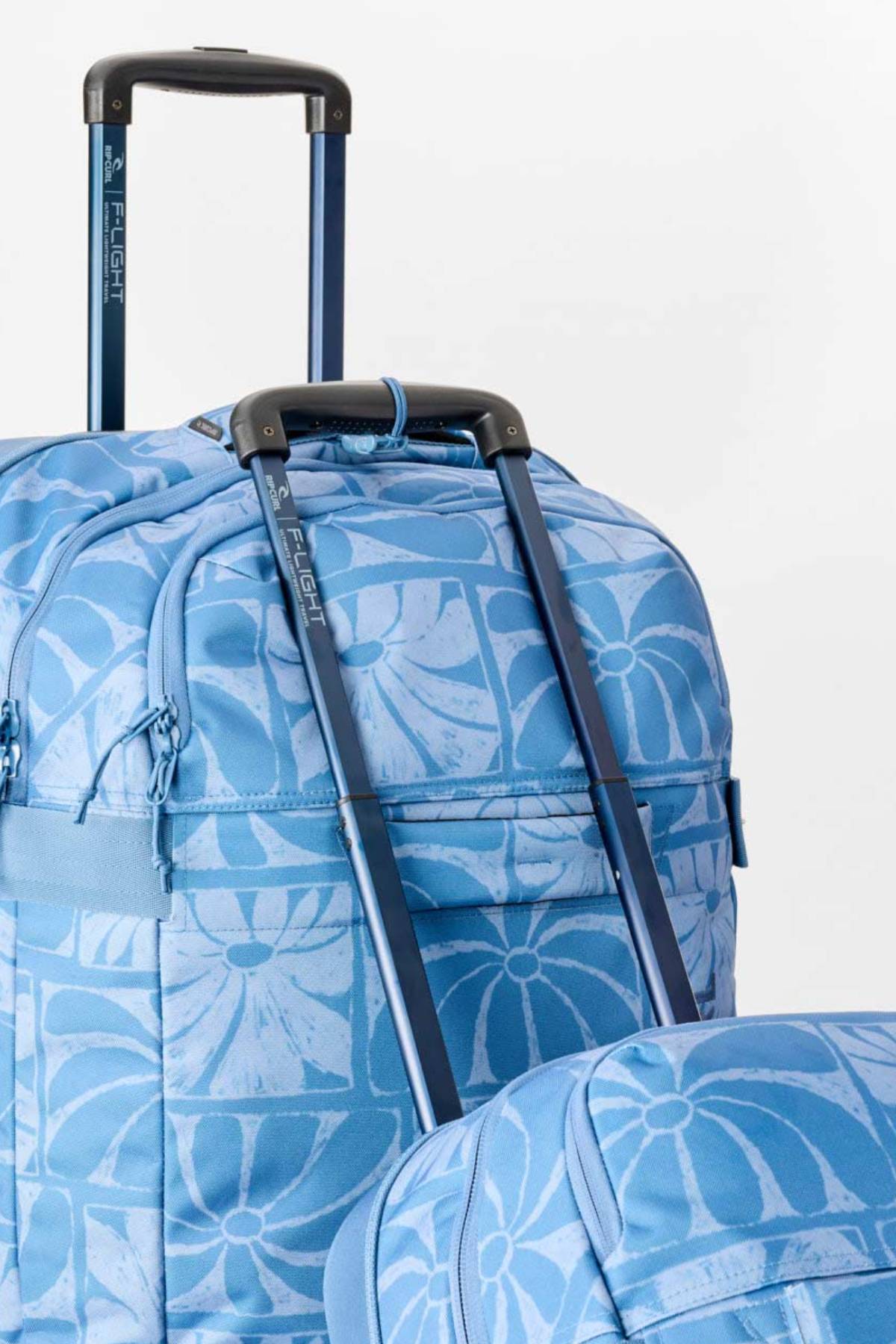Close-up of two blue Rip Curl F-Light Artist Of The Search Ty Williams 100L Global Travel Bags with floral patterns and extended telescopic handles, set against a plain white background—each spacious travel bag is perfect for your next adventure.