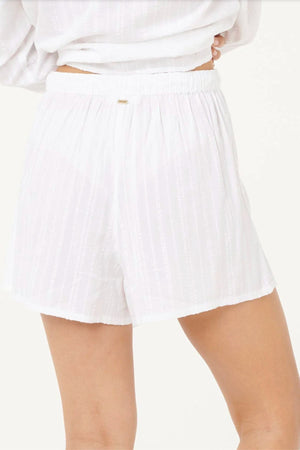 A person wears the Rip Curl Haze Short—white, loose-fitting shorts with a subtle vertical stripe and elastic waistband—perfect for casual wear. The background is light and plain.