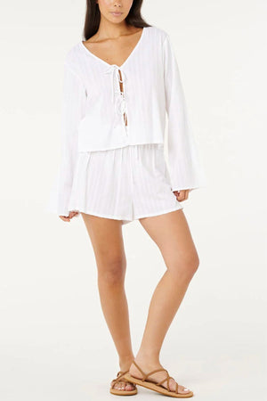 A woman wears a white long-sleeve tie-front top and Rip Curl Haze Short with brown flat sandals, standing against a plain, light background with one leg slightly bent.