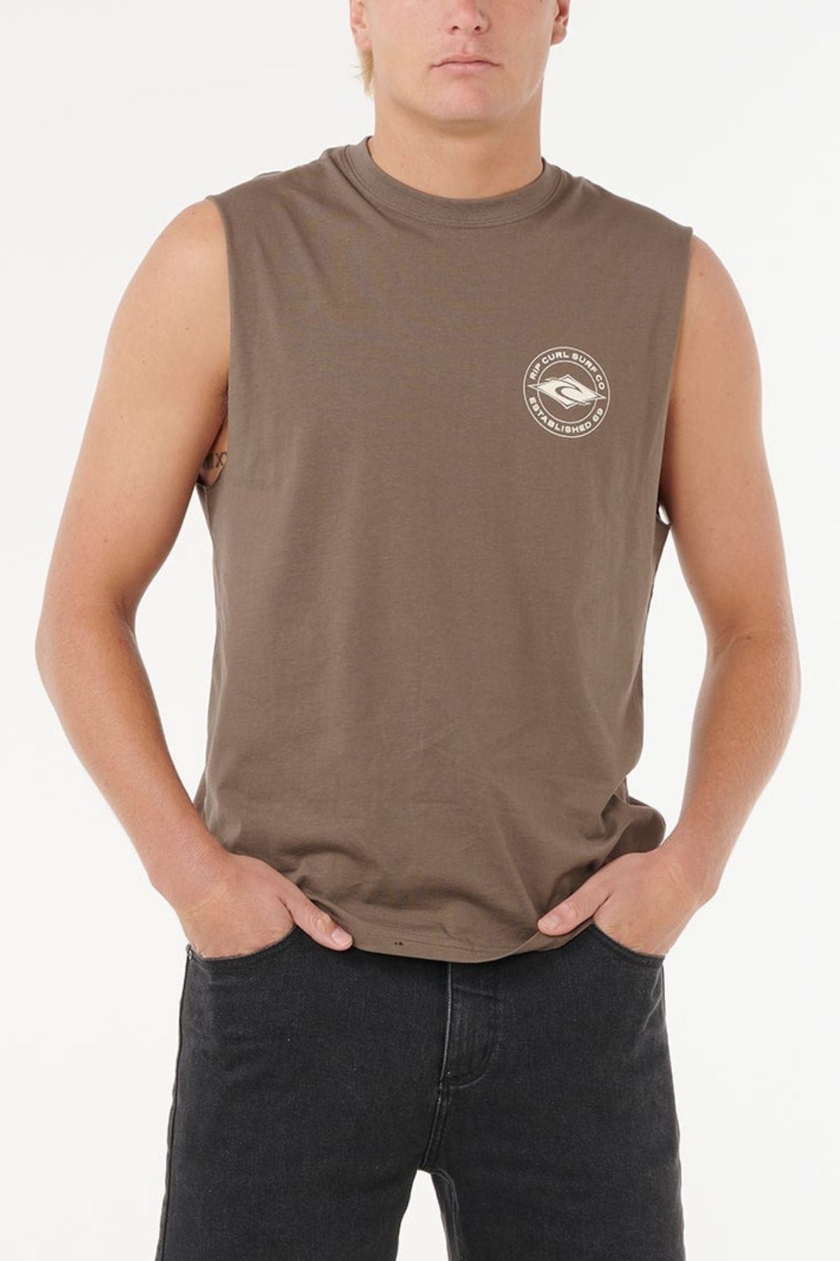 A person wears a sleeveless brown Rip Curl Staple Muscle shirt, 100% cotton with a small round chest logo, paired with black jeans, standing hands-in-pockets against a plain white background.