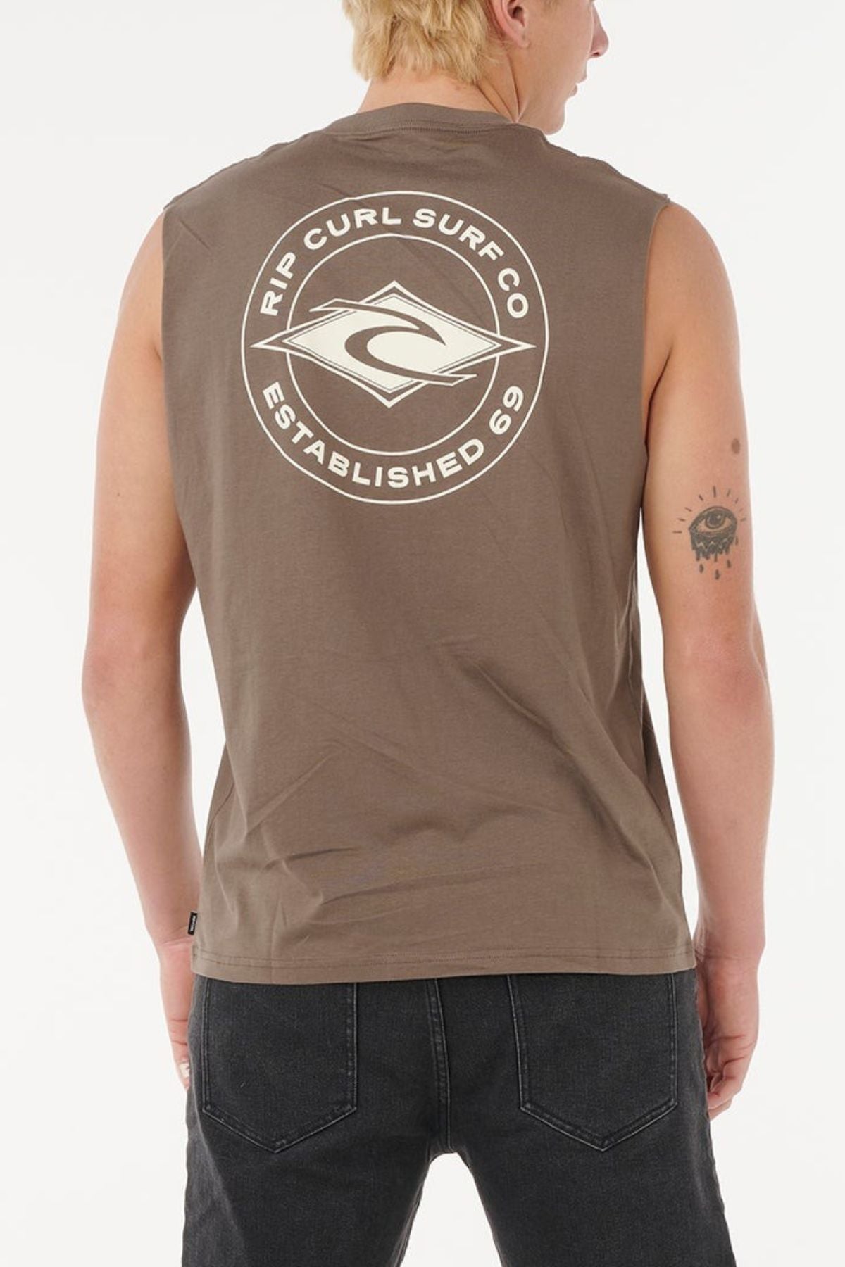 A person with short blond hair wears a brown Rip Curl Staple Muscle sleeveless shirt with a large "Rip Curl Surf Co Established 69" back logo, made from 100% cotton, paired with black jeans, and stands facing away from the camera.