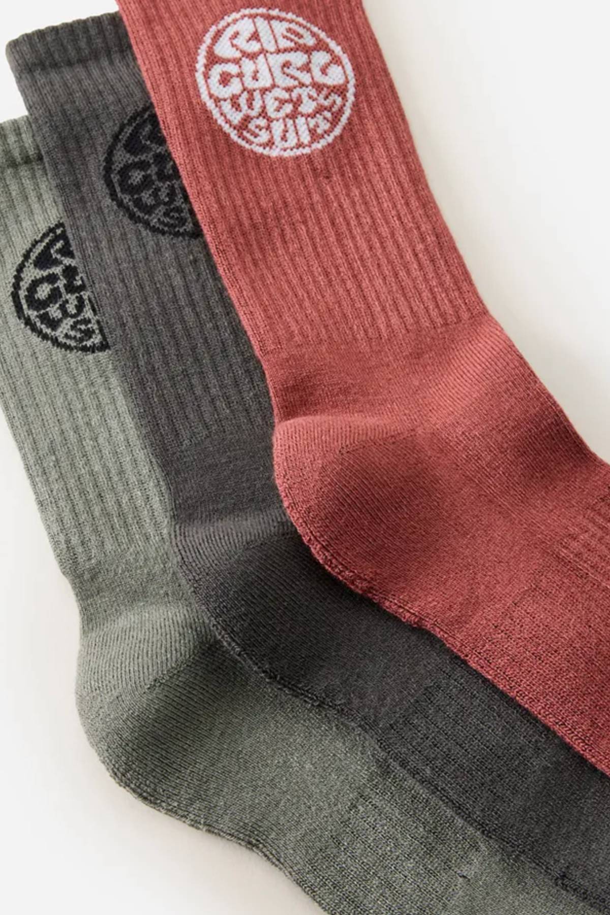 The Rip Curl Wetty Crew Sock 3 Pack includes three ribbed crew socks in olive green, dark gray, and brick red, each with a circular white "Rip Curl" logo near the top and built-in arch support for comfort.