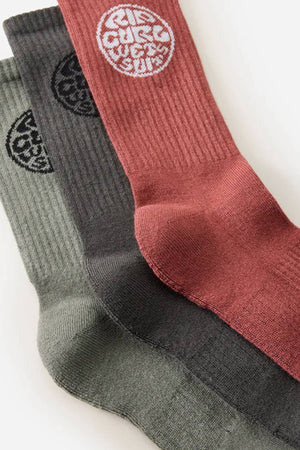The Rip Curl Wetty Crew Sock 3 Pack includes three ribbed crew socks in olive green, dark gray, and brick red, each with a circular white "Rip Curl" logo near the top and built-in arch support for comfort.