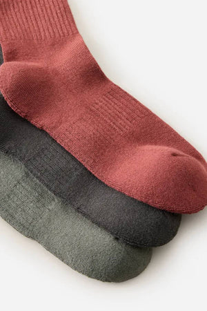 The Rip Curl Wetty Crew Sock 3 Pack features three pairs—rust red, charcoal gray, and sage green—each with comfortable arch support. The socks are neatly arranged on a light surface, overlapping one another.