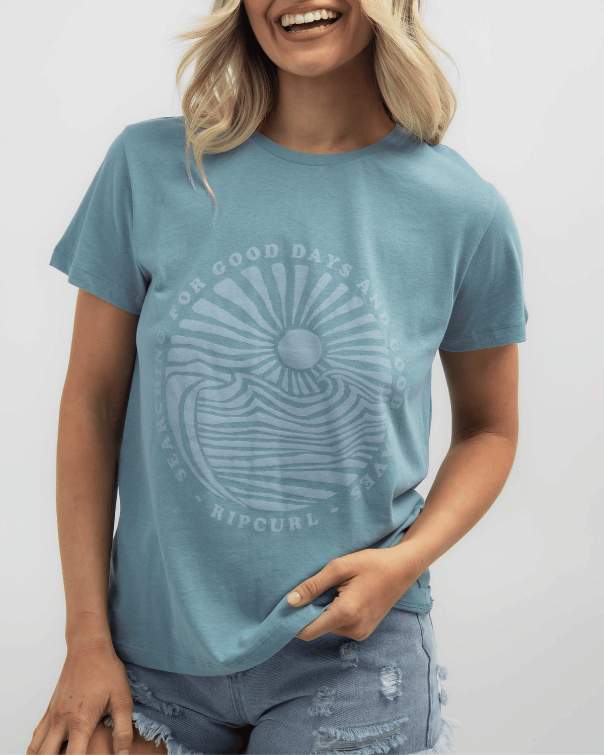 A smiling person wears the Rip Curl Good Waves Standard Tee made of cotton jersey, with a circular wave and setting sun design. Text reads "Searching for Good Days and Good Waves, Ripcurl." They pair it with distressed denim shorts.