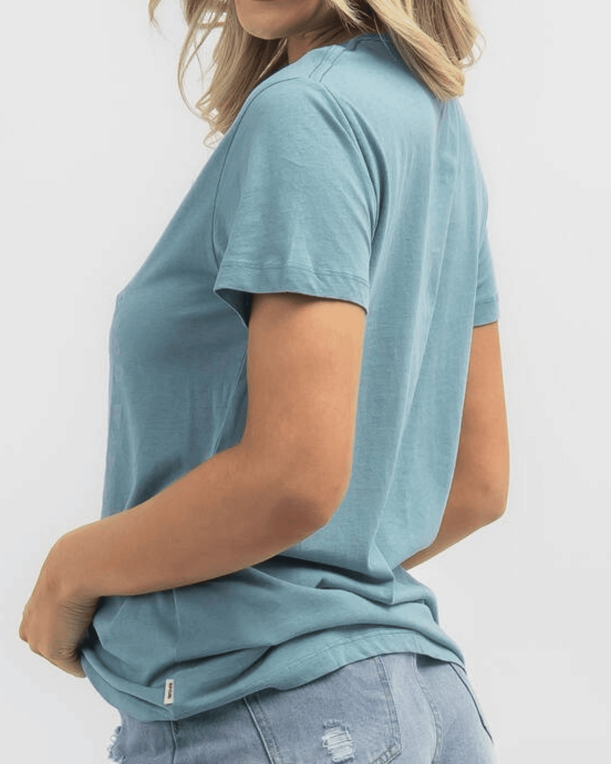 A person in a blue Rip Curl Good Waves Standard Tee and distressed denim shorts drapes comfortably, with one hand resting in the back pocket, against a plain white backdrop.