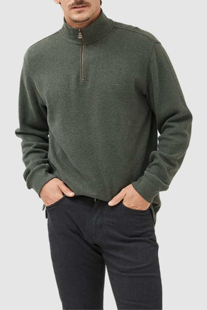 A person is wearing a dark green Rodd & Gunn Alton Ave Sweat, made of 100% cotton with a high collar. They stand with their hands in pockets, against a light gray background, pairing it with black pants while their face remains partially visible.