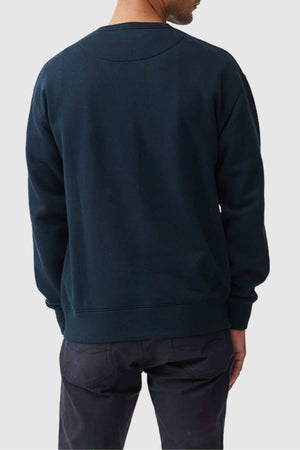 A person is standing with their back to the camera, wearing a dark blue Rodd & Gunn Gunn Logo Sweat featuring embroidered logo and dark pants. The background is plain and light-colored. The person's head is turned slightly to the right, but their face is not visible.