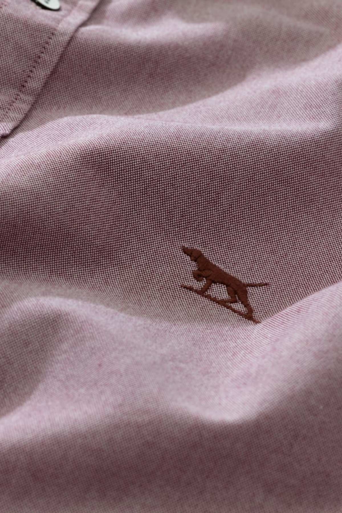 Close-up of the Rodd & Gunn Gunn Oxford Sports Fit Shirt in 100% cotton mauve, highlighted by a small embroidered brown dog in a running pose, visible stitching, and partial top button for a refined look.