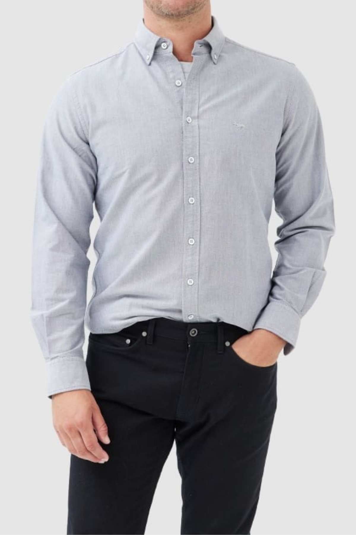 A person standing against a neutral background, wearing a stylish Rodd & Gunn Gunn Oxford Sports Fit Shirt in light gray. The shirt is made of 100% cotton and features long sleeves, paired with black pants. Their left hand is in their pants pocket while the right hand rests by their side. Their head is not visible in the image.