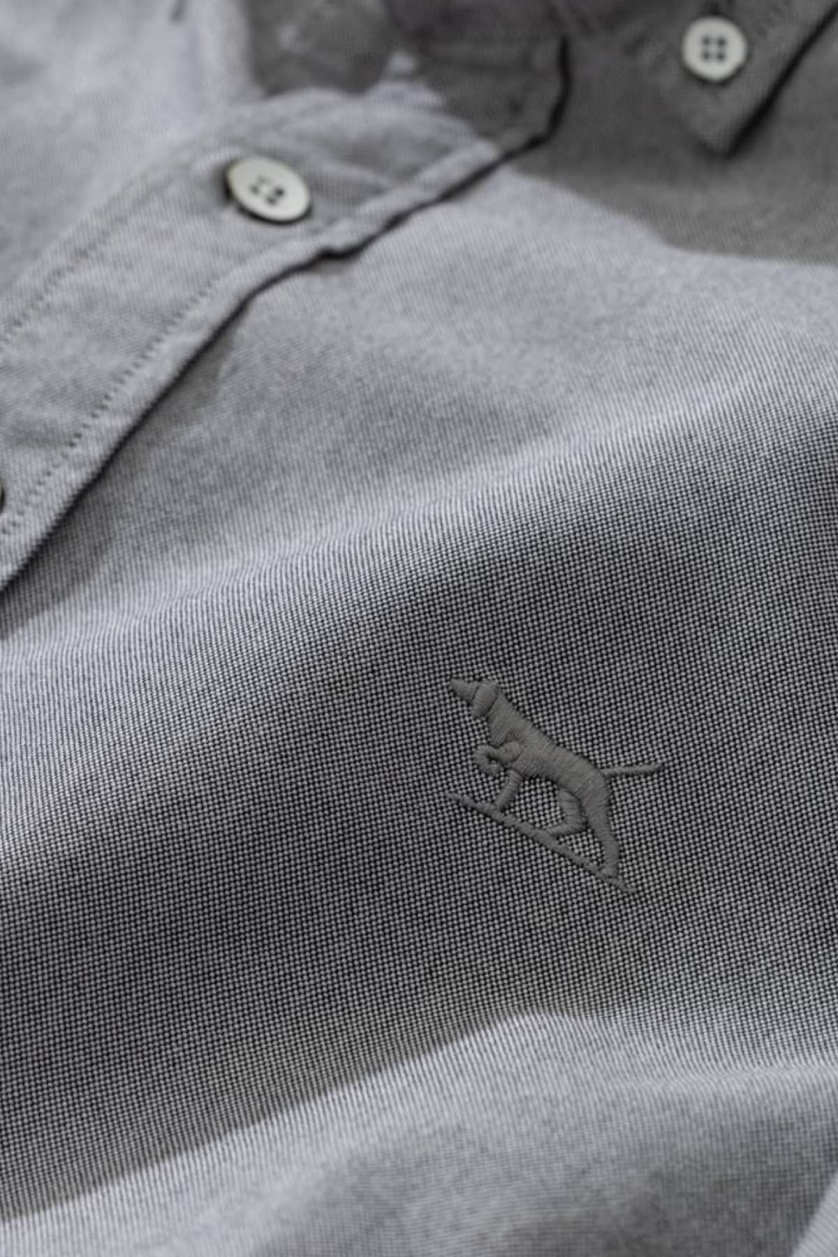 A close-up of the Rodd & Gunn Gunn Oxford Sports Fit Shirt showcases its stylish grey button-up design, crafted from 100% cotton. The fabric exhibits a fine texture, and the chest features an intricately embroidered logo of a dog in motion. In the background, the collar and two white buttons are partially visible.