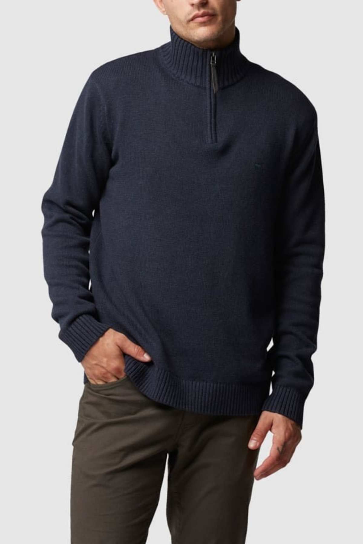 A person is dressed in a Rodd & Gunn Merrick Bay Knit sweater in dark navy, featuring long sleeves and a premium YKK half-zipper at the collar, paired with olive green pants. Made of 100% cotton, they have their right hand in their pocket and are posing against a plain, light gray background.