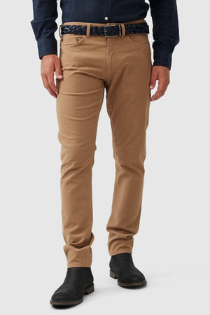 A person stands against a plain background, wearing Rodd & Gunn Motion 2 Straight Jeans in dark blue comfort stretch fabric, paired with black boots. A black braided belt complements the YKK zip fly, with only the lower half of the body shown.