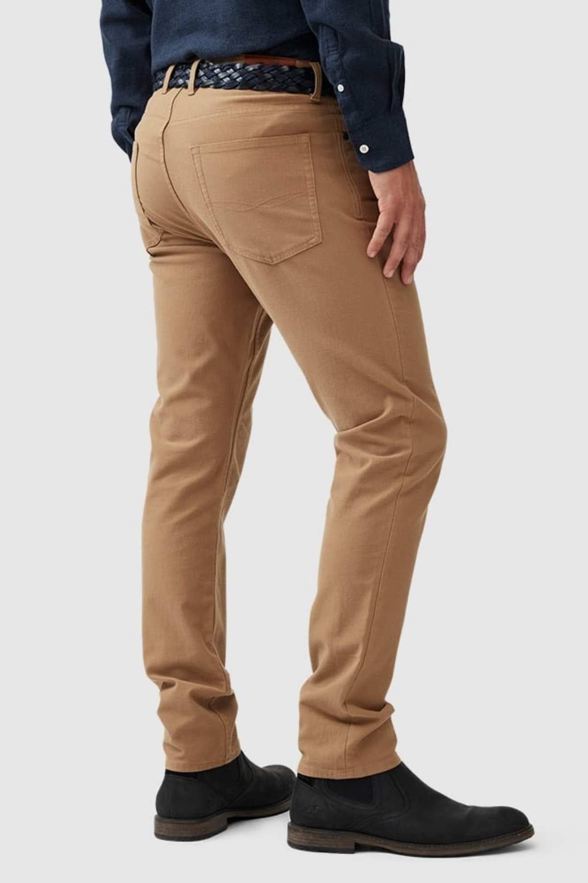 Wearing Rodd & Gunn Motion 2 Straight Jeans with a YKK zip fly, tan pants, and a dark blue shirt, the person faces away. Their outfit includes black suede boots and a woven belt against a plain, light-colored background.