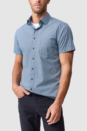 A man wears the Rodd & Gunn Chartwell Sports Fit Shirt, a slim fit, short-sleeved blue patterned 100% cotton shirt with a front pocket, paired with dark pants, standing against a plain light gray background.