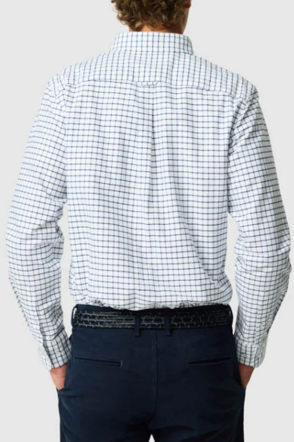 A person with curly hair, seen from behind, wears the Rodd & Gunn Gunn Check Oxford 3 Sports Fit Shirt in long sleeves and dark pants with a black belt, hands in pockets.