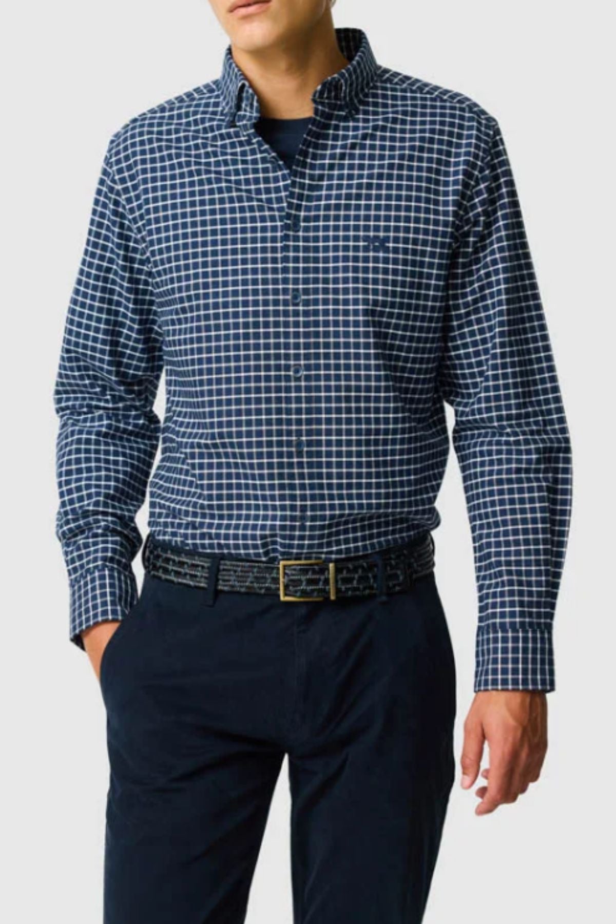 A man wears the Rodd & Gunn Gunn Check Oxford 3 Sports Fit Shirt, a long-sleeve blue and white check Australian cotton style, tucked into dark navy trousers with a black belt. The plain light gray background and his head are partially out of frame.