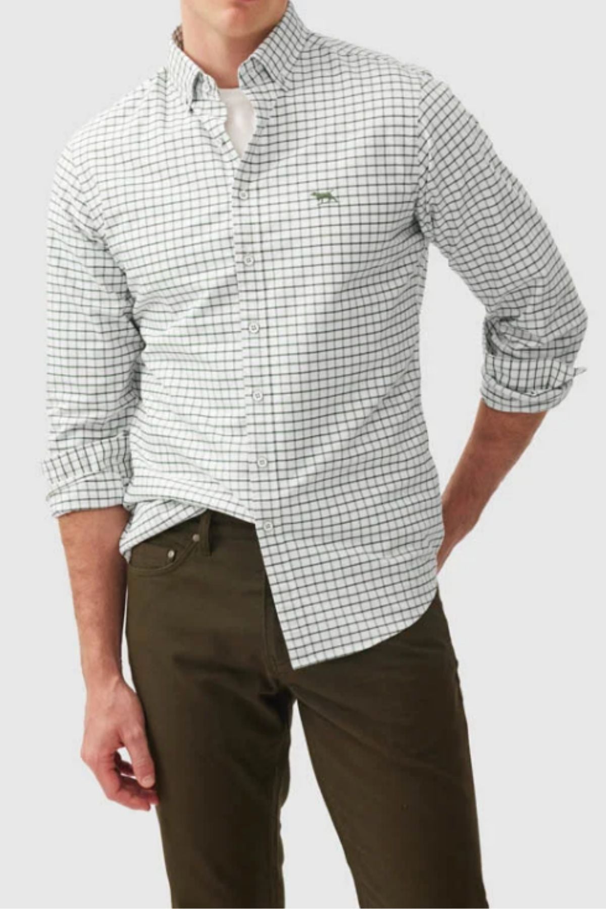 A man wears the Rodd & Gunn Gunn Check Oxford 3 Sports Fit Shirt in white with a black grid pattern and rolled-up sleeves, paired with dark olive green pants, standing with one hand in his pocket.