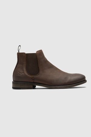 The Rodd & Gunn Ealing Chelsea Boot features brown oiled leather, elastic side panels, a heel pull tab, low stacked heel, and flexible rubber outsole. Shown on a plain light gray background.