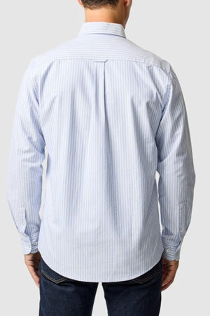 A man, seen from the back against a plain light gray background, wears the Rodd & Gunn Gunn Oxford Stripe 4 Of Shirt in light blue and white vertical stripes with dark jeans.