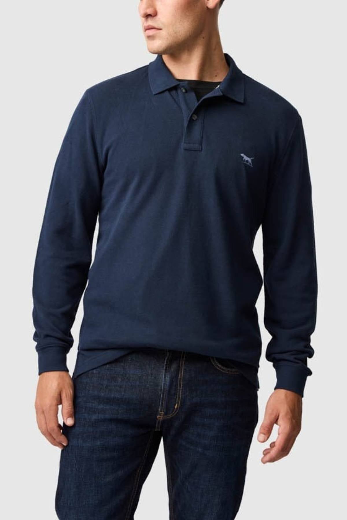 A man wearing a Rodd & Gunn Long Sleeve Sports Fit Gunn Polo in navy, made from breathable pique mesh fabric, and dark blue jeans, stands against a light gray background.