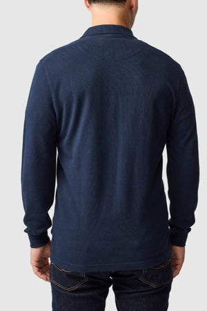 A man in a Rodd & Gunn Long Sleeve Sports Fit Gunn Polo (dark blue, 100% cotton pique mesh) and dark jeans stands facing away from the camera against a plain light gray background.