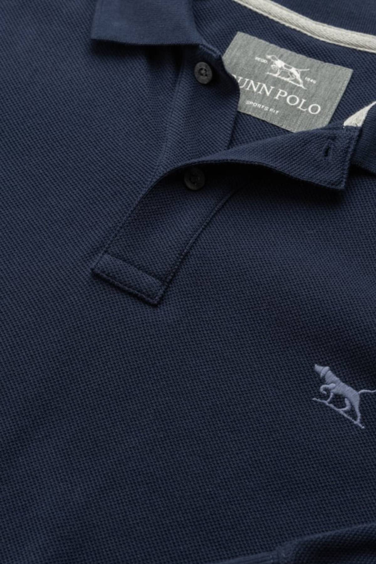 A close-up of the Rodd & Gunn Long Sleeve Sports Fit Gunn Polo in navy blue, featuring 100% cotton breathable pique mesh, two buttons, a collar, and a small embroidered dog logo on the chest. The inside label reads "DUNN POLO.