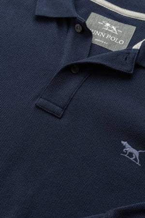 A close-up of the Rodd & Gunn Long Sleeve Sports Fit Gunn Polo in navy blue, featuring 100% cotton breathable pique mesh, two buttons, a collar, and a small embroidered dog logo on the chest. The inside label reads "DUNN POLO.