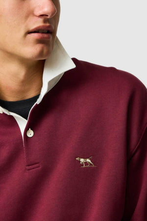 Wearing the Rodd & Gunn Otago Sweat, a maroon rugby collar shirt with white buttons and an embroidered dog logo, a person stands against a plain light background. Only their lower face and upper torso are shown.