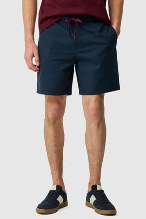 A person in a burgundy shirt, navy Rodd & Gunn Resort Short with an elastic waist, and navy sneakers featuring white accents stands with hands in pockets against a plain background.