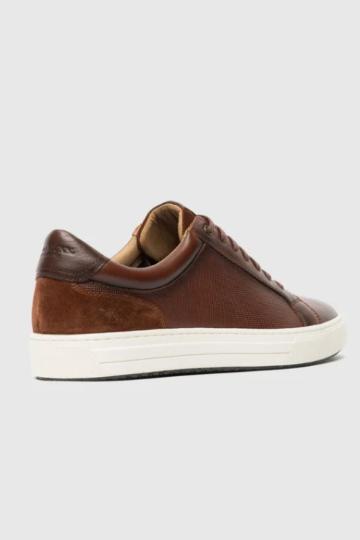 The Rodd & Gunn Transit Sneaker is a brown leather and suede low-top with a white rubber sole, shown from the back-left against a plain light gray background.