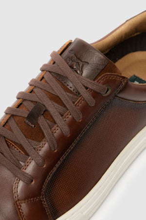 Close-up of the Rodd & Gunn Transit Sneaker: brown leather with textured side panel, brown laces, and white rubber sole. Embossed logo on tongue and visible stitching showcase its premium craftsmanship.