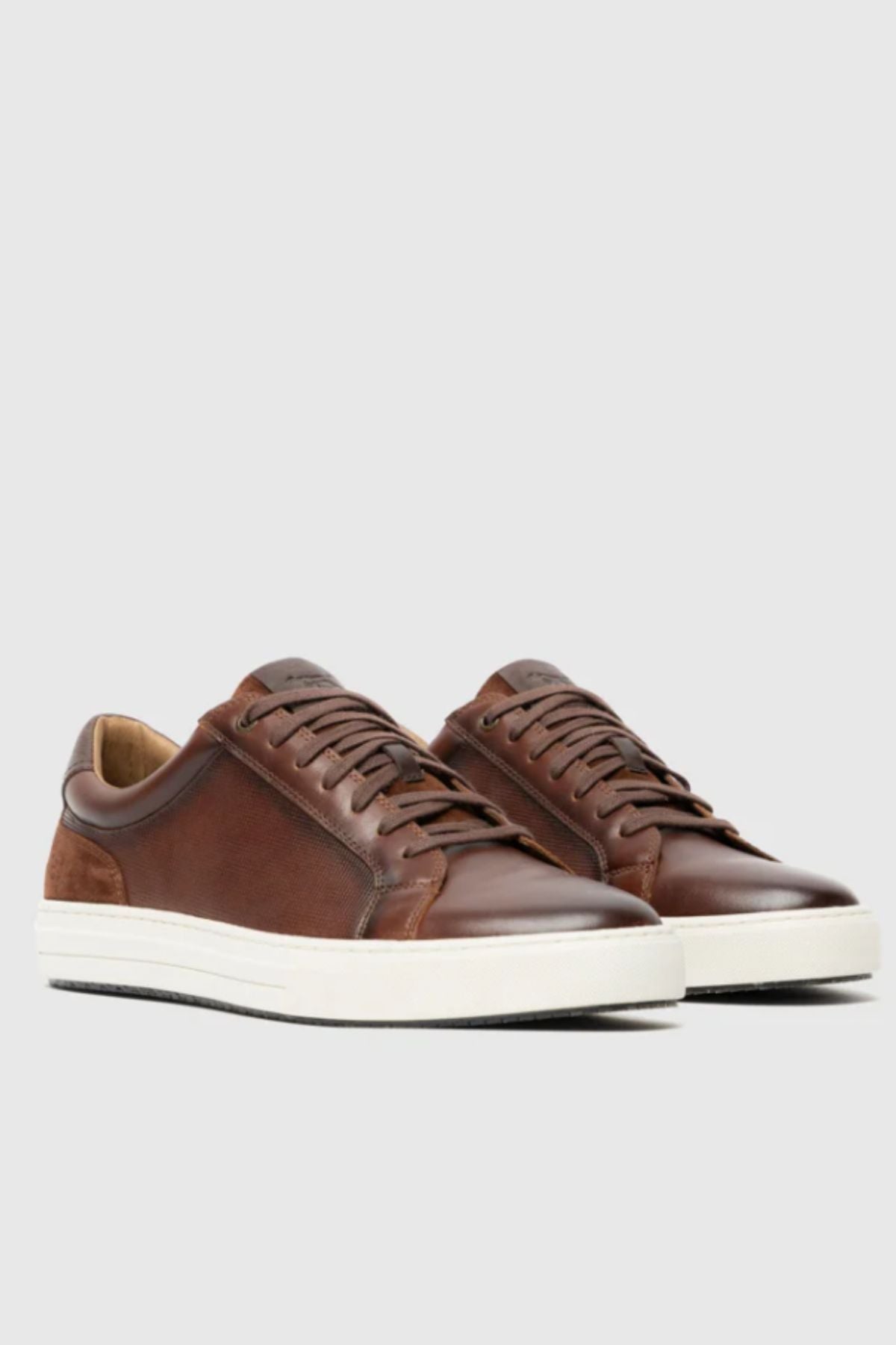 A pair of Rodd & Gunn Transit Sneakers in brown leather with white rubber soles, brown laces, and suede heel accents are displayed on a plain light gray background.