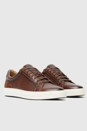 A pair of Rodd & Gunn Transit Sneakers in brown leather with white rubber soles, brown laces, and suede heel accents are displayed on a plain light gray background.