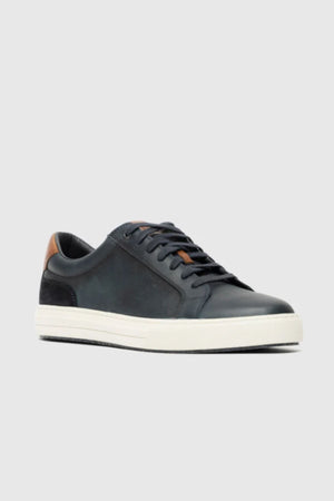 The Rodd & Gunn Transit Sneaker is a black low-top leather sneaker with black laces, a white rubber sole, and a brown heel accent, shown against a plain light gray background.