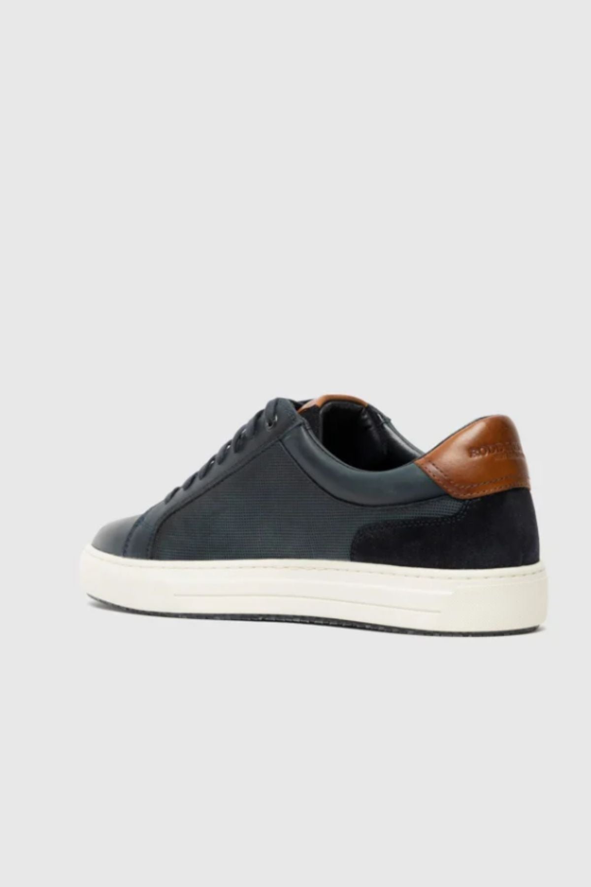 The Rodd & Gunn Transit Sneaker is a black low-top leather shoe with a white sole, black laces, and a brown leather heel accent, displayed at an angle on a plain light gray background.
