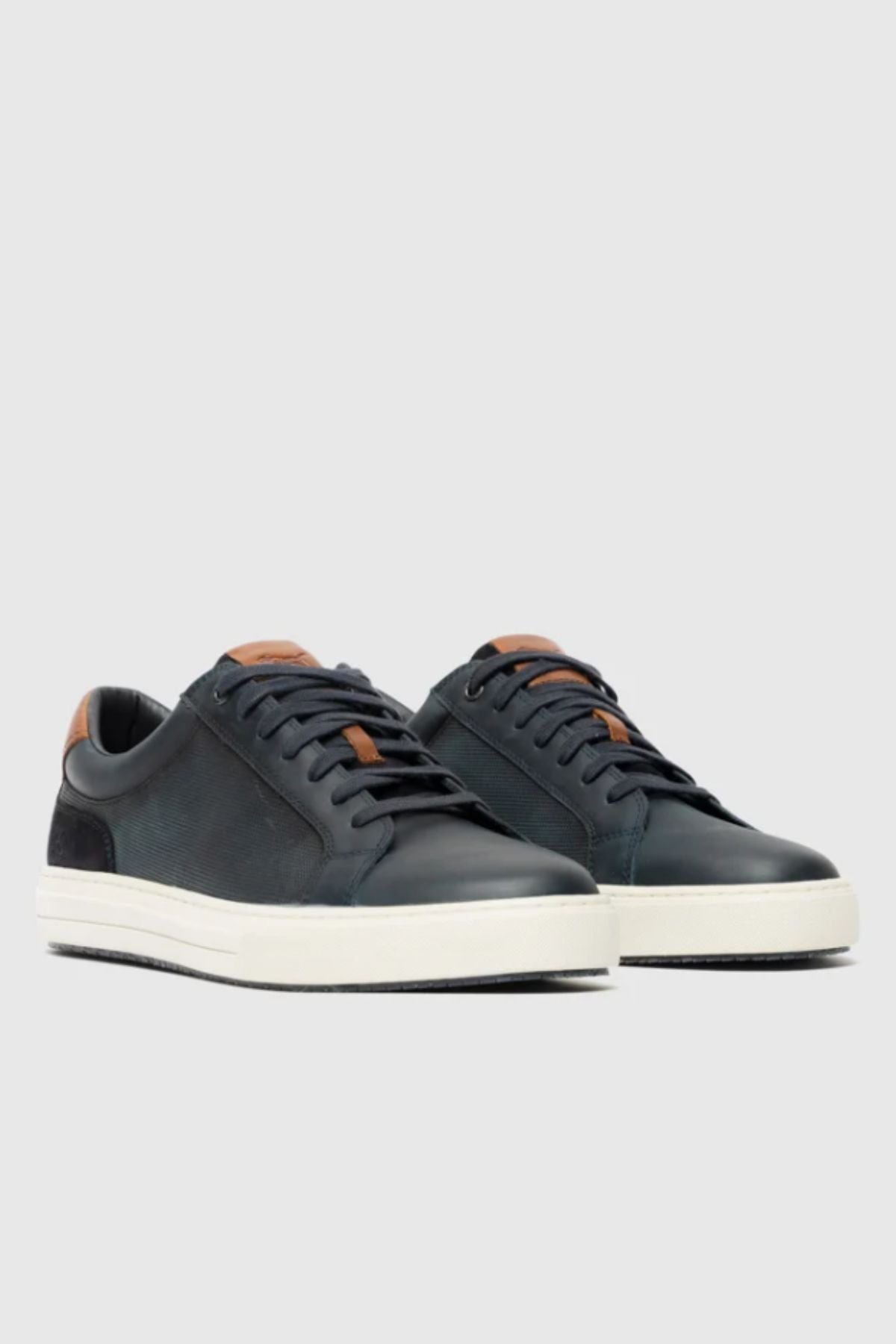 A pair of Rodd & Gunn Transit Sneakers in black with white soles, black laces, and tan leather accents on the tongue and heel are displayed on a light gray background.